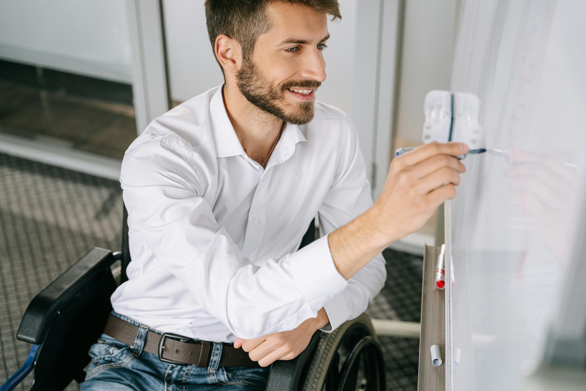 A cheerful man in a wheelchair writing ideas on a whiteboard indoors.