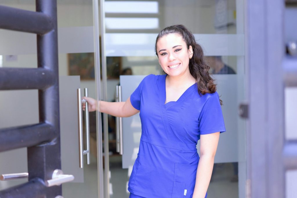 Smiling female nurse in blue scrubs opens a glass door in a healthcare setting.